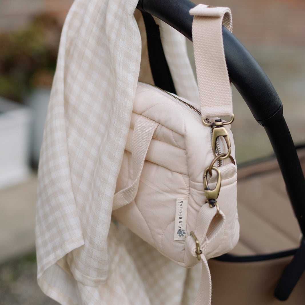 Beige baby carrier with a quilted cover draped over it, set against a blurred outdoor background.