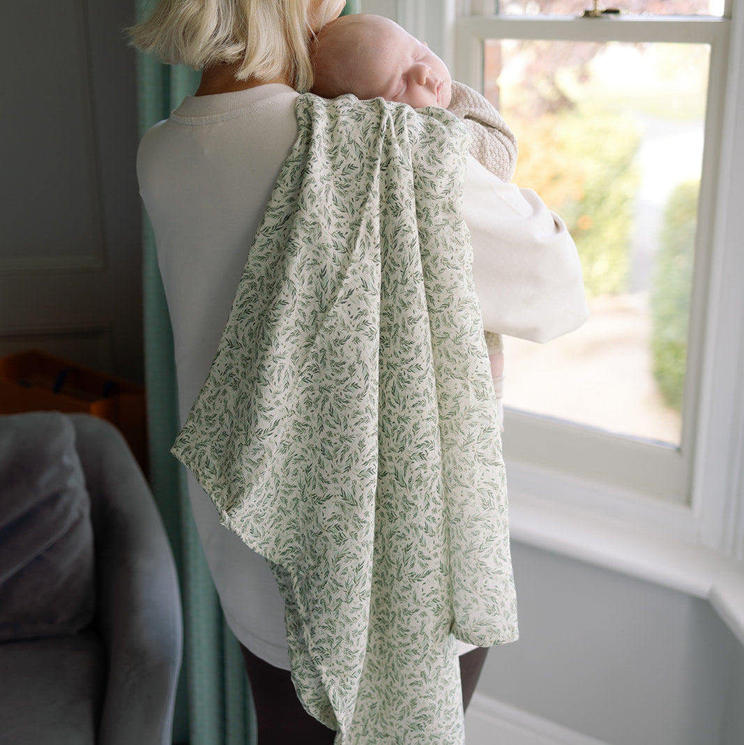 Person holding a baby wrapped in a green floral blanket by a window.