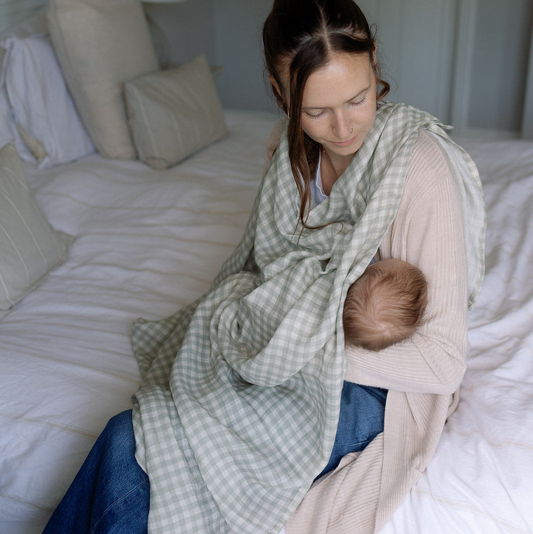 Woman sitting on a bed holding a baby wrapped in a green checkered wrap.