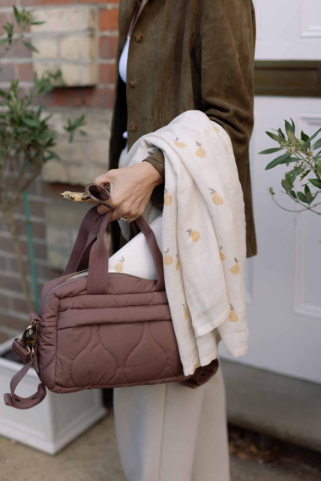 Person holding a brown quilted handbag with a white coat draped over their arm, standing outdoors.