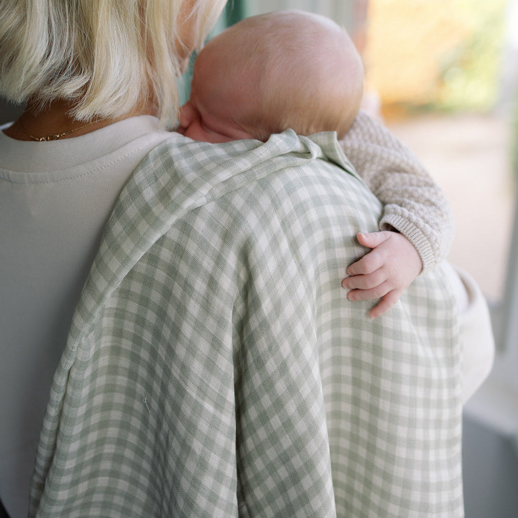 Woman holding a baby wrapped in a green checkered blanket indoors.