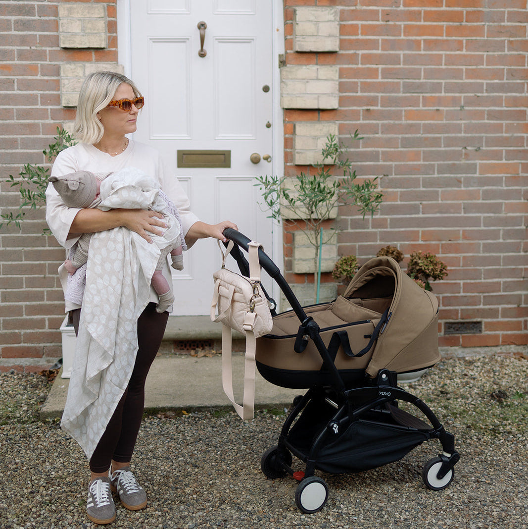 Woman holding a baby and pushing a stroller in front of a brick building with a white door.