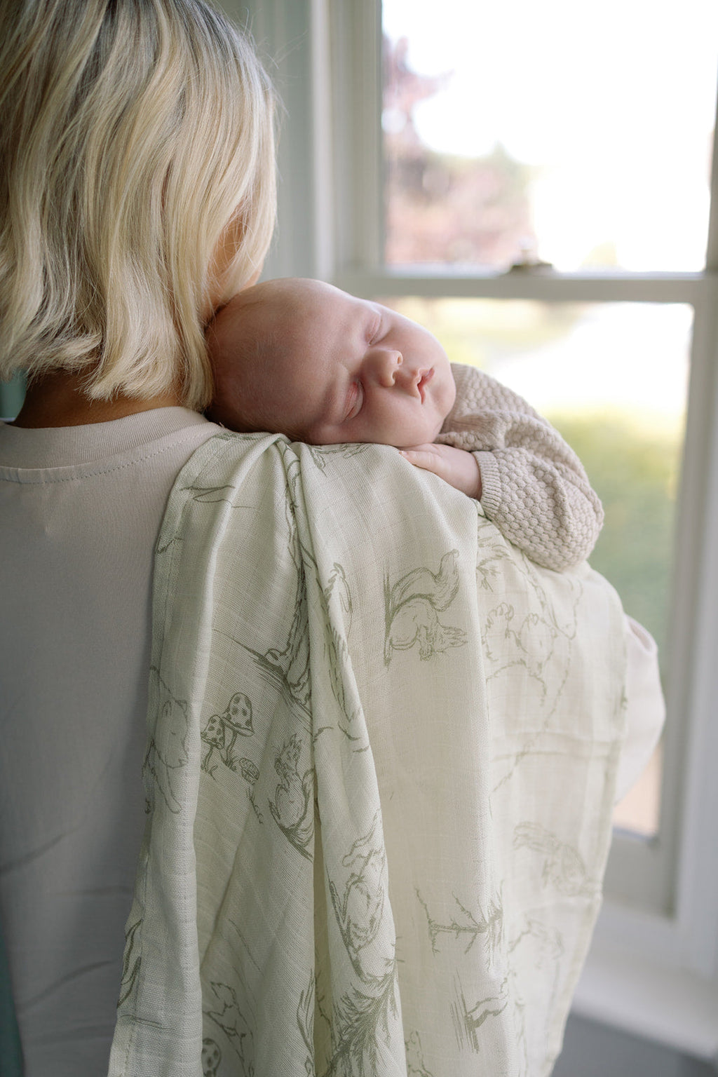 Person holding a sleeping baby wrapped in a floral-patterned blanket by a window.
