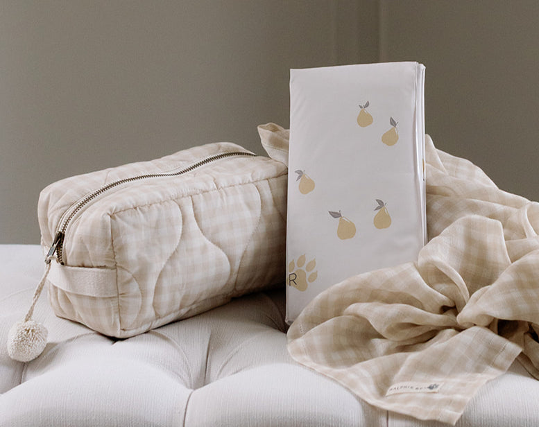 Beige quilted bag, white box with fruit design, and beige blanket on a light gray surface.