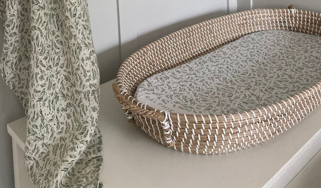 Woven pet bed with patterned cushion on a white surface next to a floral curtain.