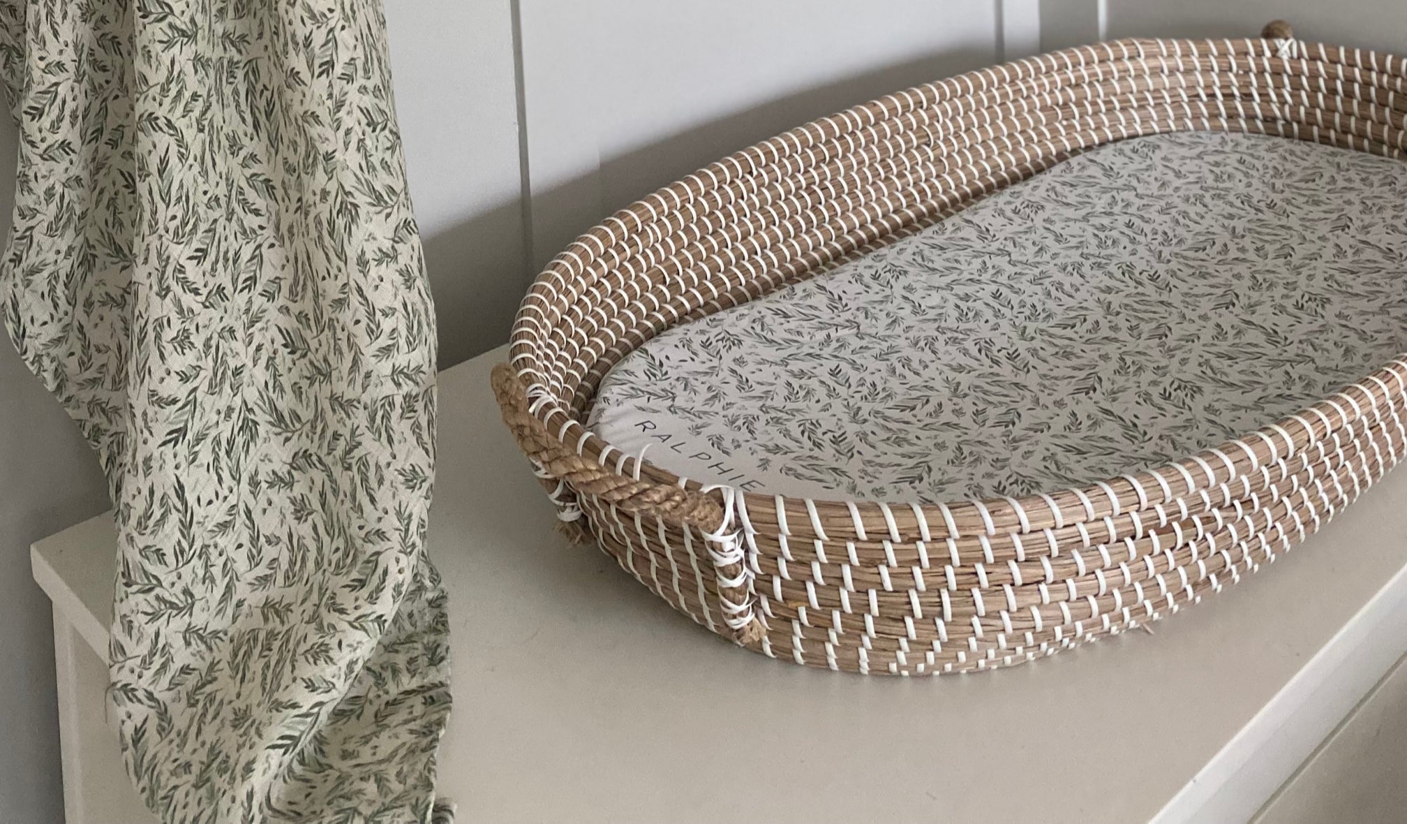 Woven pet bed with patterned cushion on a white surface next to a floral curtain.
