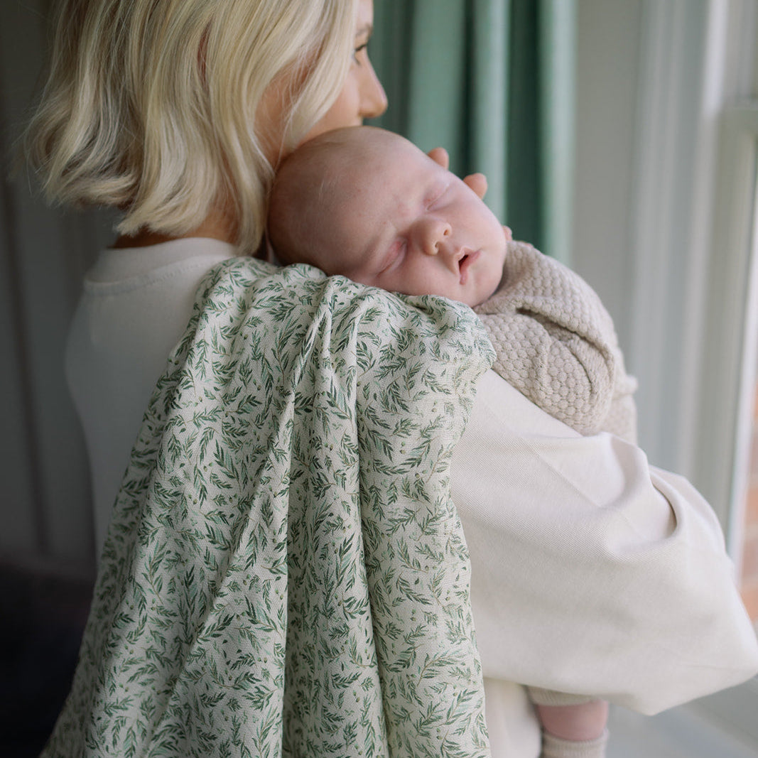 Woman holding a baby wrapped in a green floral blanket by a window.