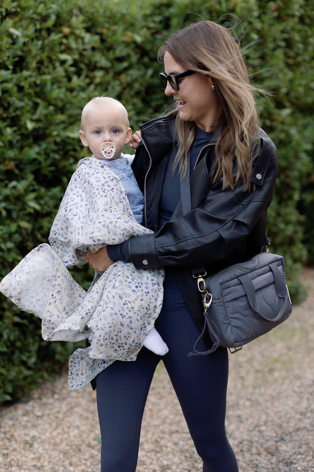 Woman holding a baby wrapped in a blanket outdoors with greenery in the background