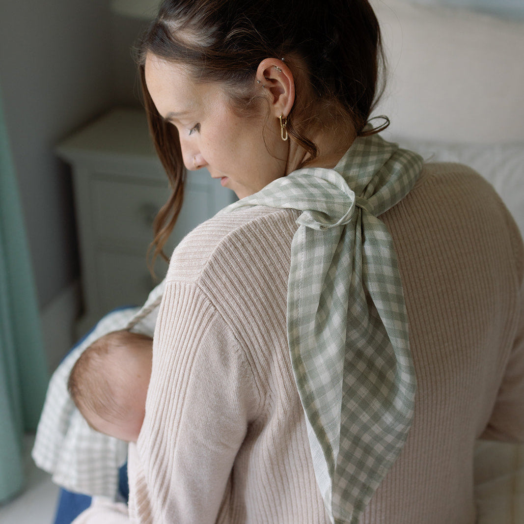 Woman sitting on a bed wearing a beige sweater with a green checkered scarf over her shoulder.