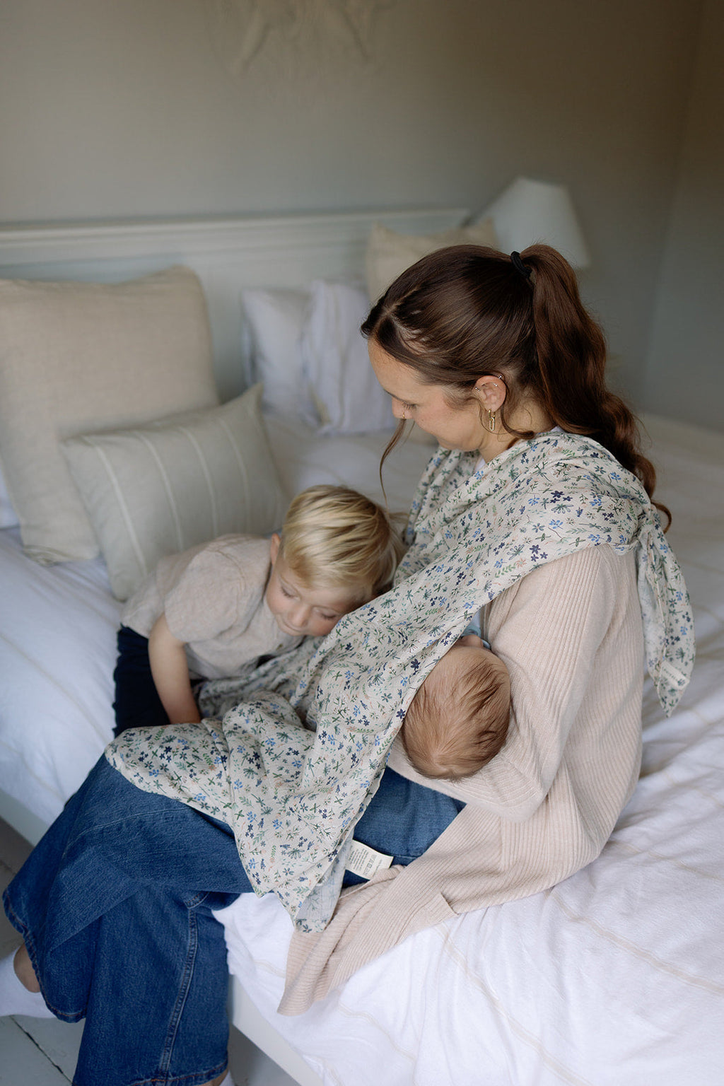 Woman sitting on a bed with two children, one of whom is being held and the other lying next to her.