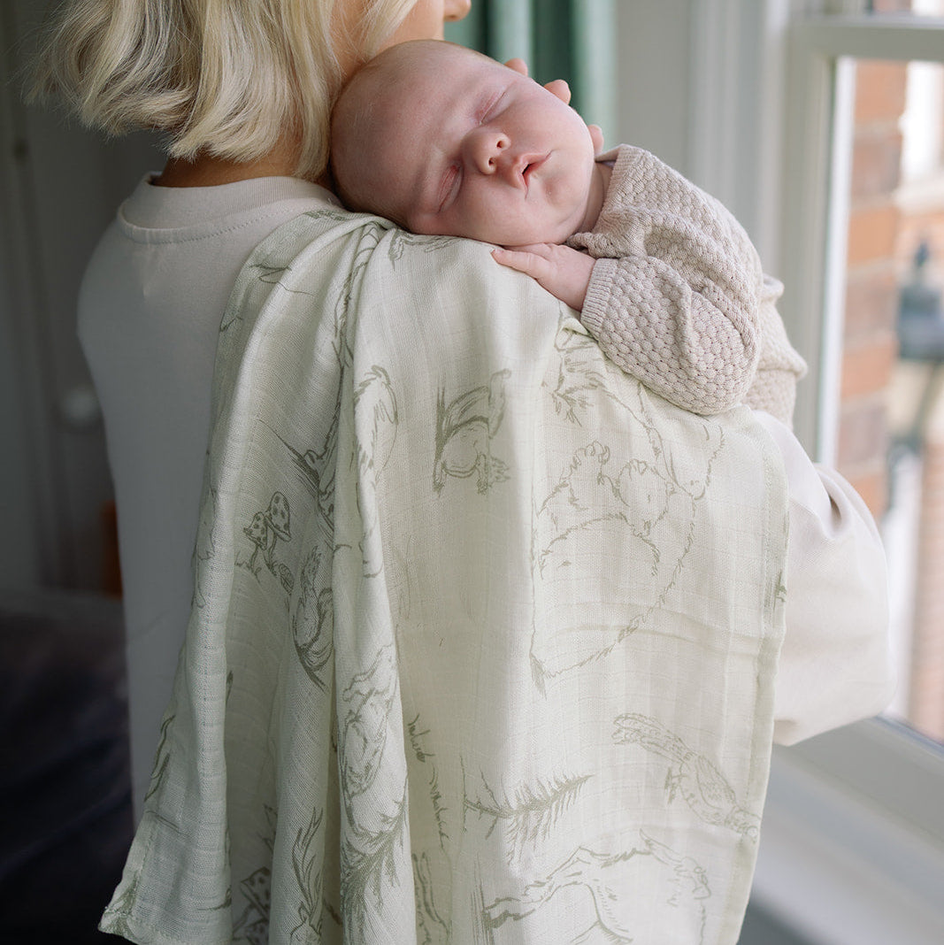 Woman holding a baby wrapped in a light green blanket with floral patterns near a window.