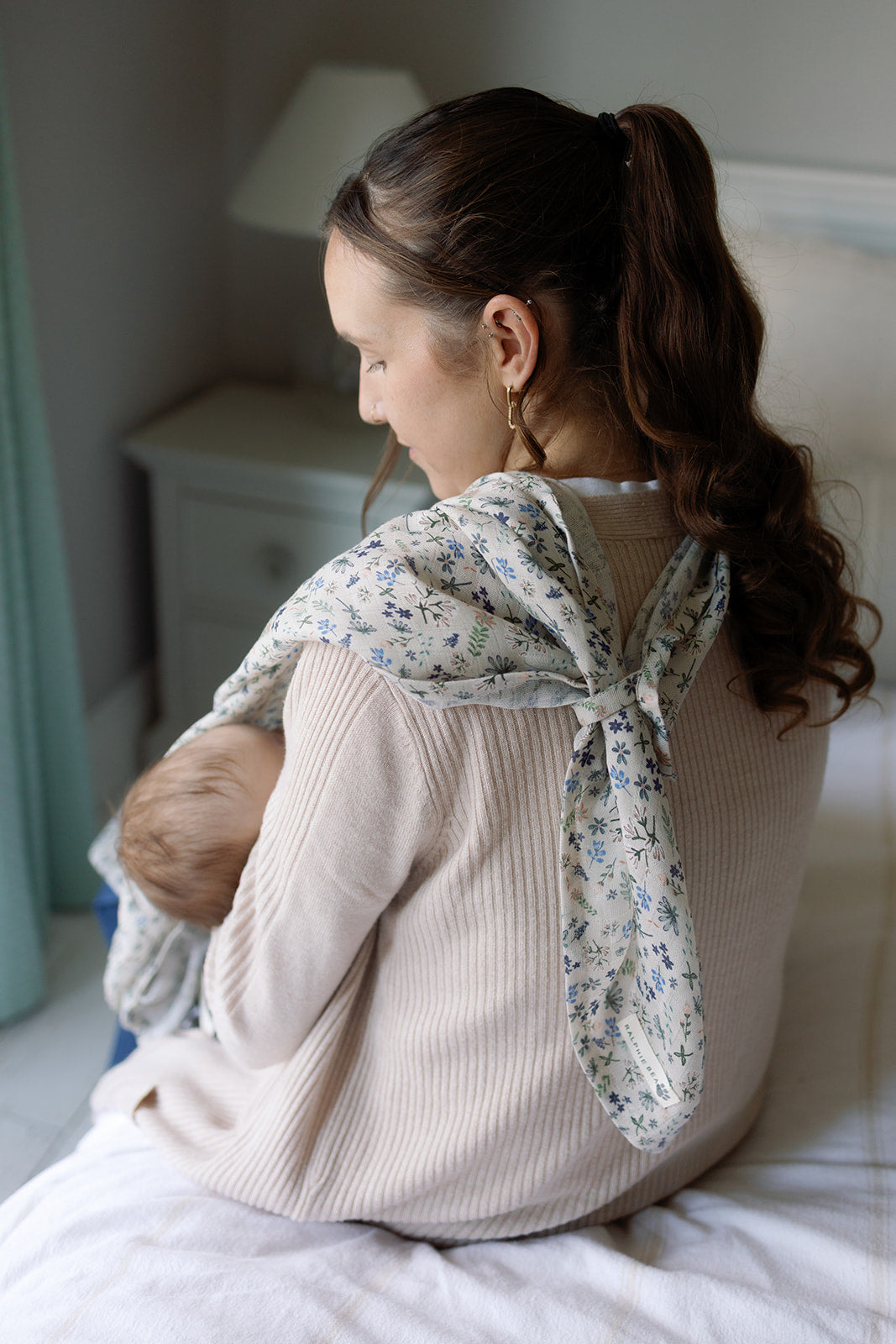 Woman sitting on a bed wearing a floral scarf, with a baby in a sling.