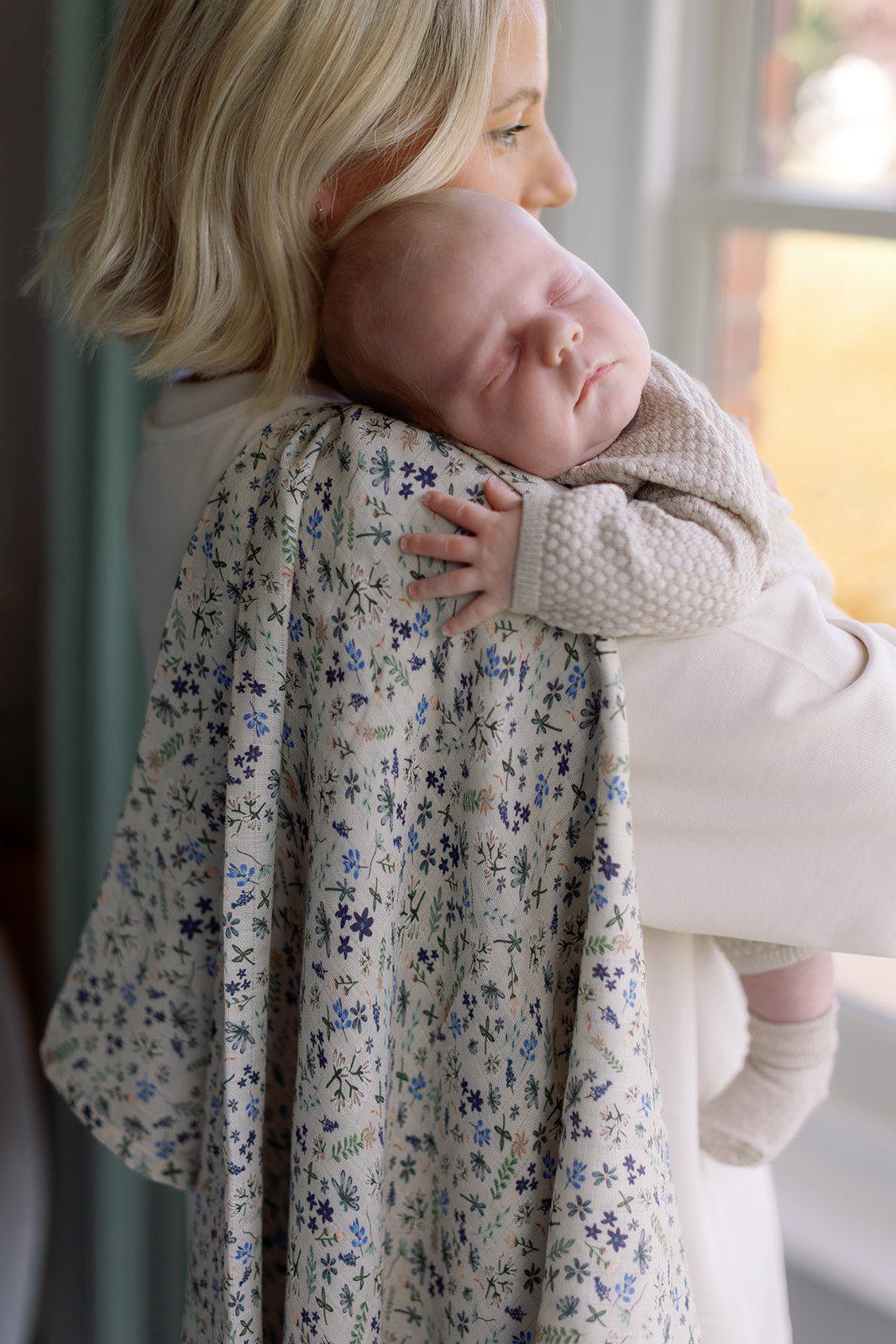 Woman holding a baby wrapped in a floral blanket indoors.