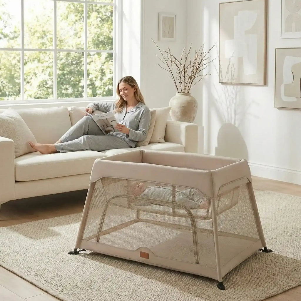 Woman reading a book on a couch with a baby bassinet next to her in a bright living room.