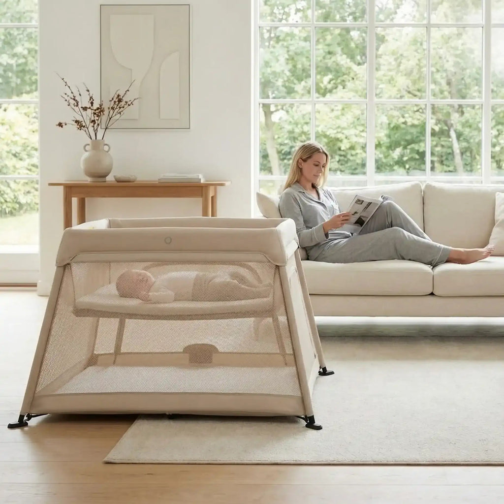Woman reading a book on a couch with a baby in a portable crib in a bright living room.