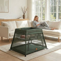 Green baby crib in a living room with a woman reading a book on a couch.
