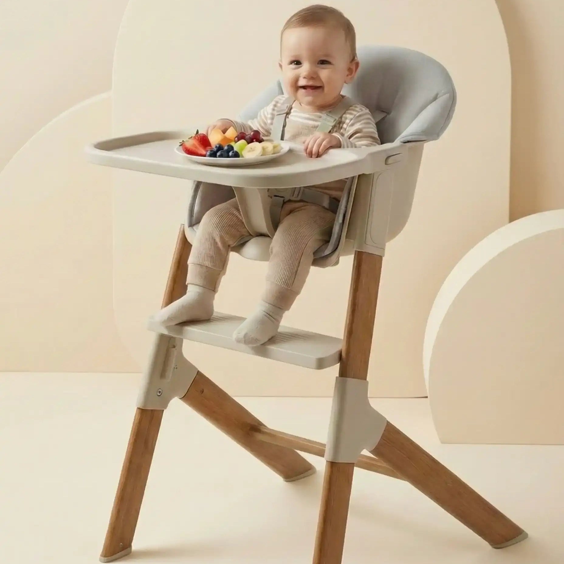Baby sitting in a modern high chair with a tray of fruits on a neutral background
