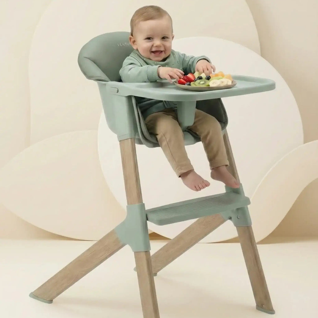 Child sitting in a green and beige high chair with a plate of food on a white background