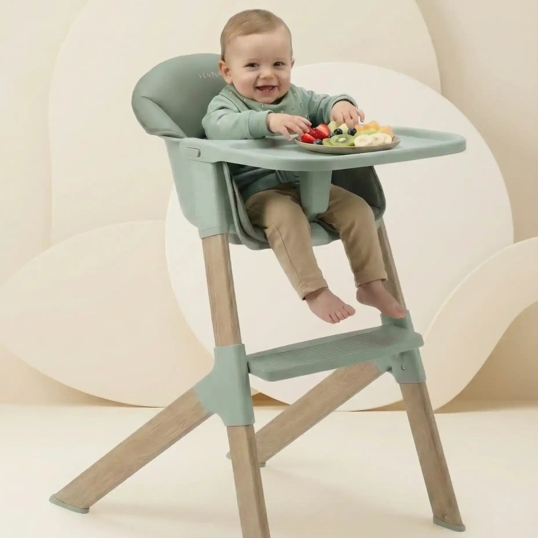 Child sitting in a green and beige high chair with a plate of food on a white background