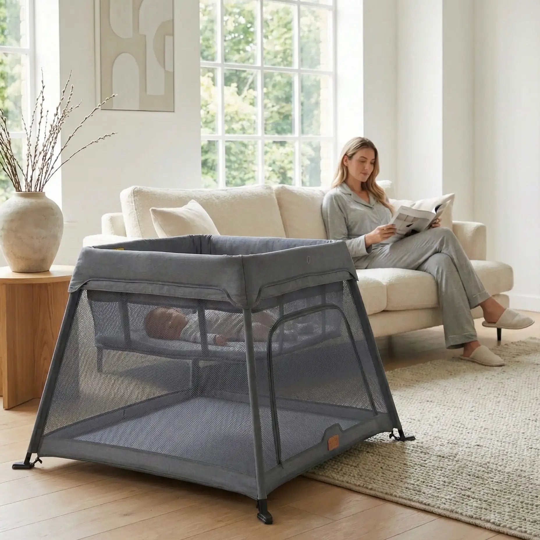 Portable crib in a living room with a woman reading a newspaper on a couch.