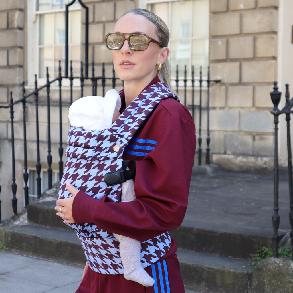 Person wearing sunglasses and a maroon tracksuit with a baby in a houndstooth-patterned carrier outdoors.