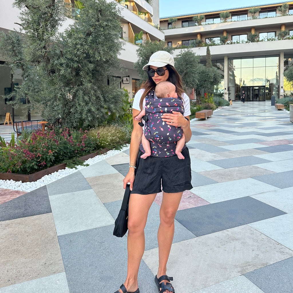 Woman carrying a baby in a sling while standing on a checkered pavement with modern buildings in the background.