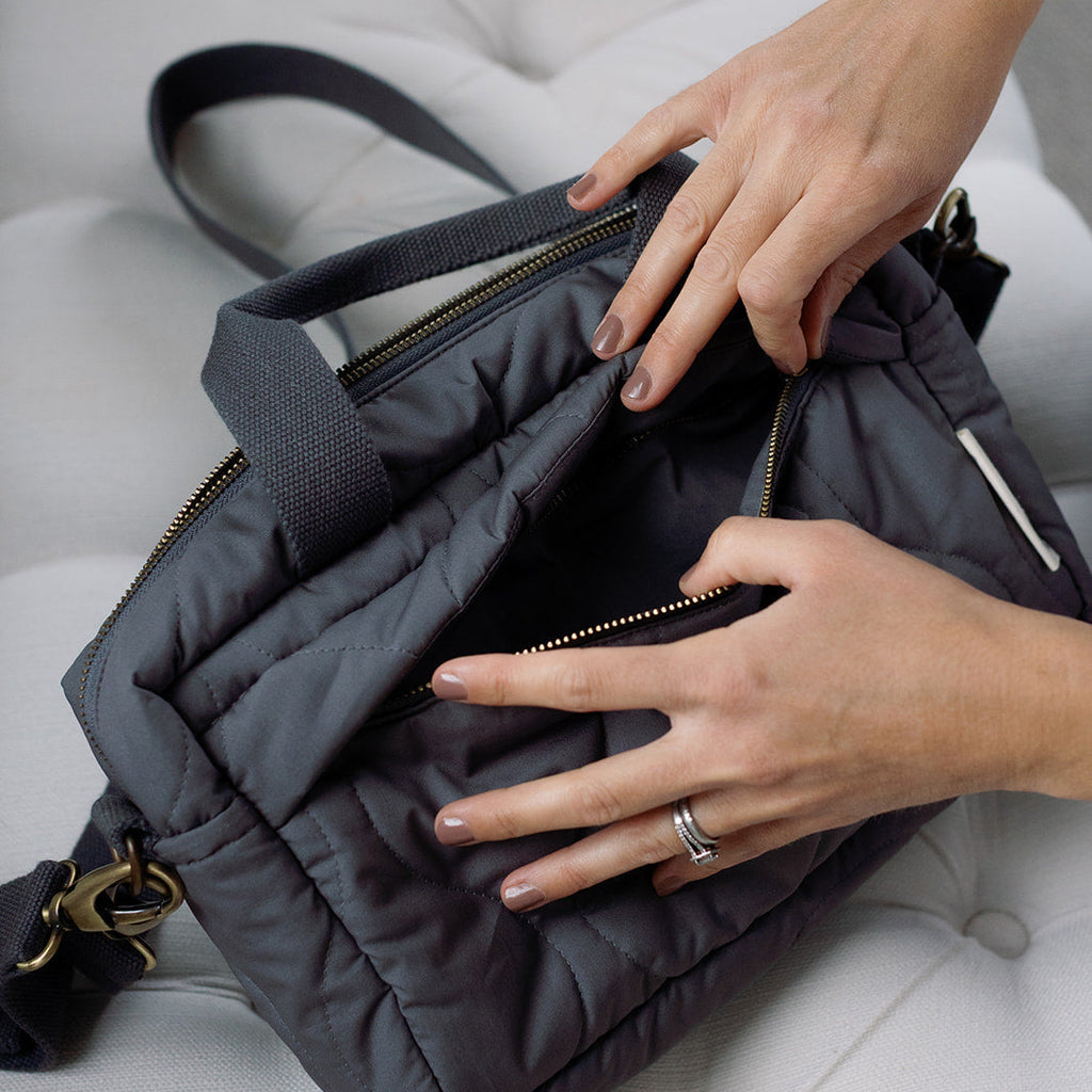 Person opening a dark gray handbag on a light gray surface