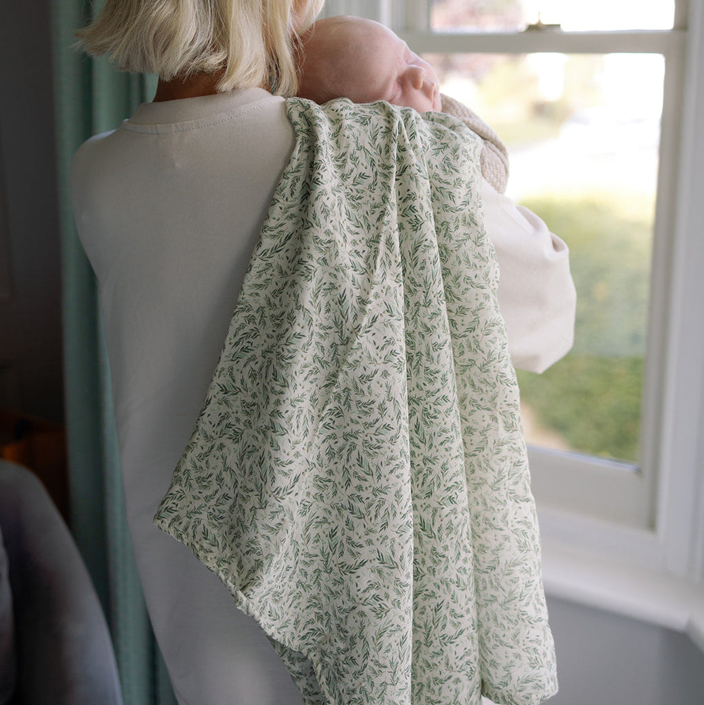 Woman holding a baby wrapped in a green patterned blanket by a window.