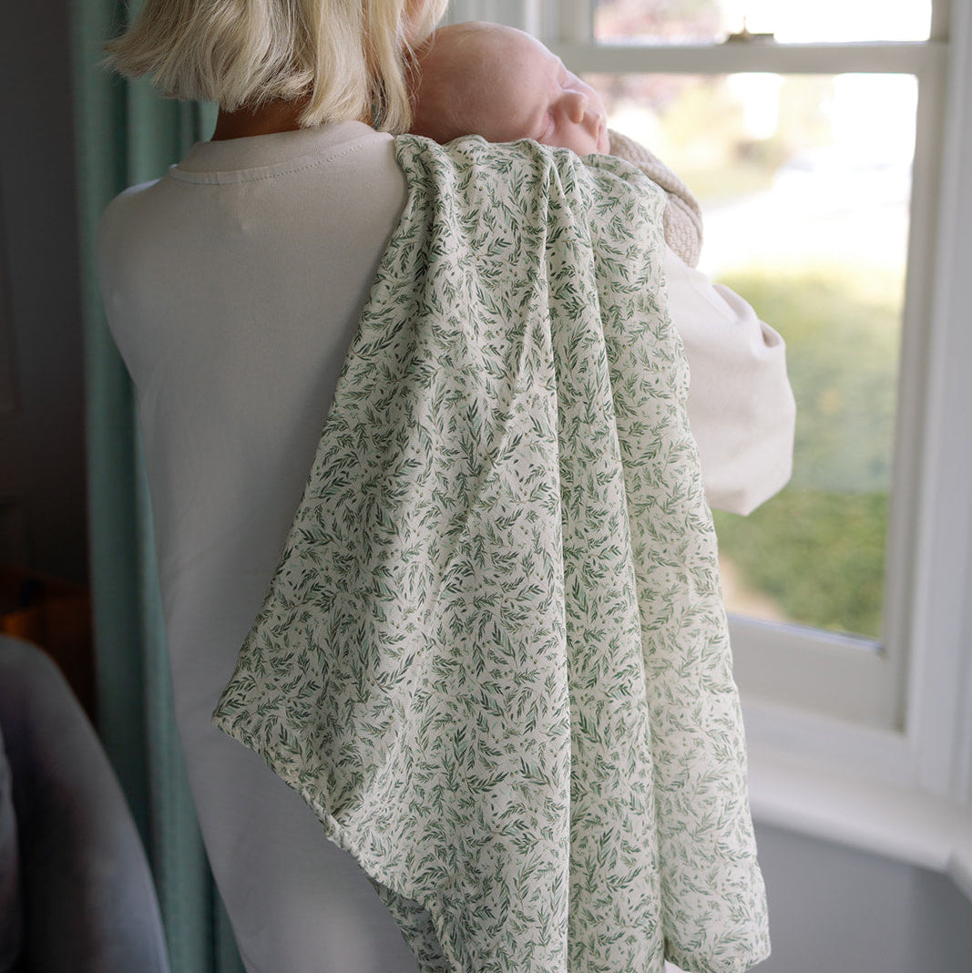 Woman holding a baby wrapped in a green patterned blanket by a window.