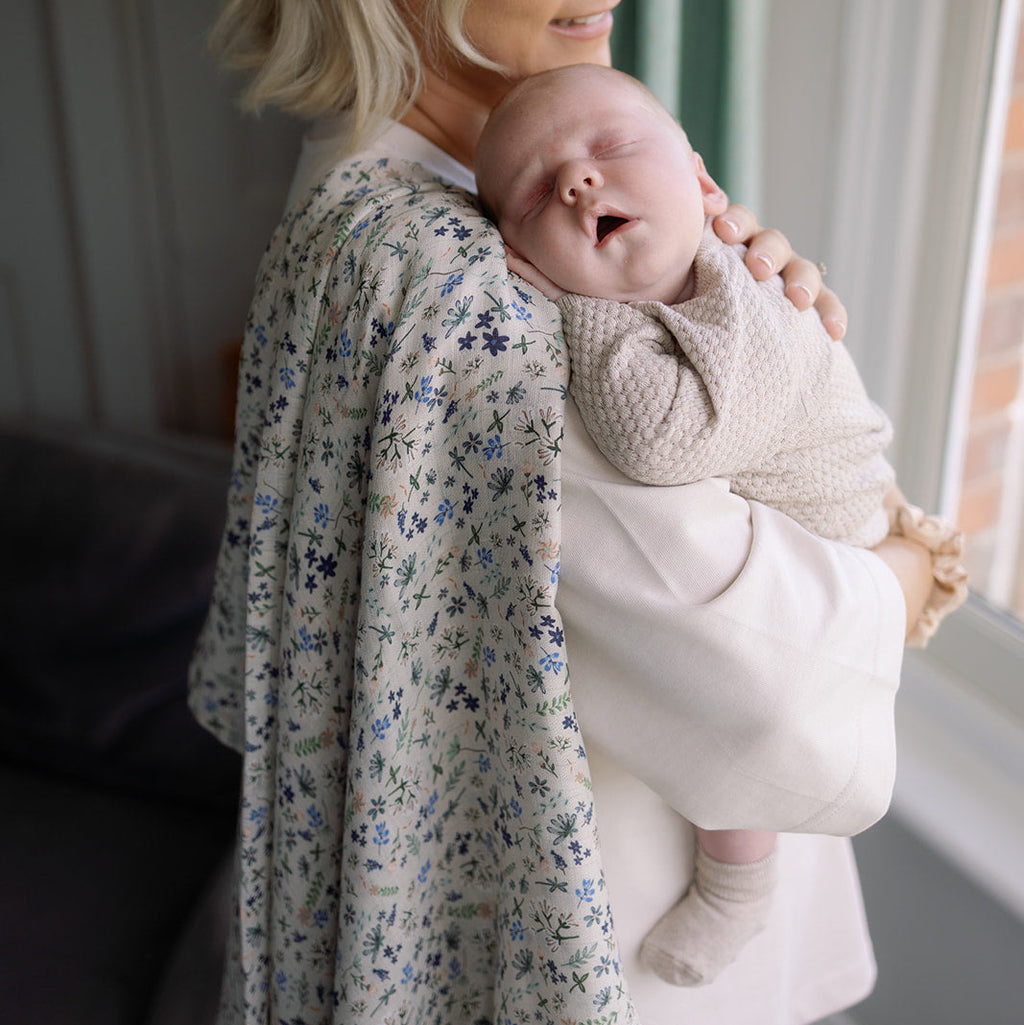 Woman holding a baby in a room with a window and green curtain.
