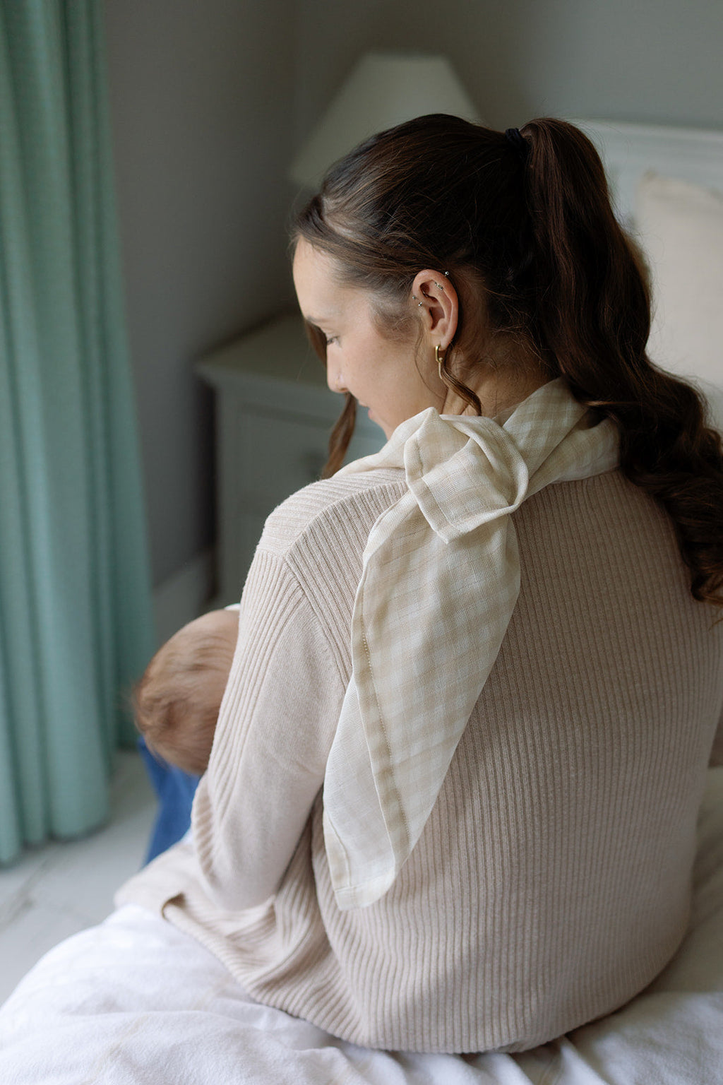 Woman sitting on a bed wearing a beige sweater with a white scarf, looking out of frame.